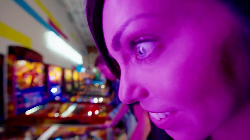 A woman plays a pinball machine bathed in vivid purple light with a row of others glowing in the background