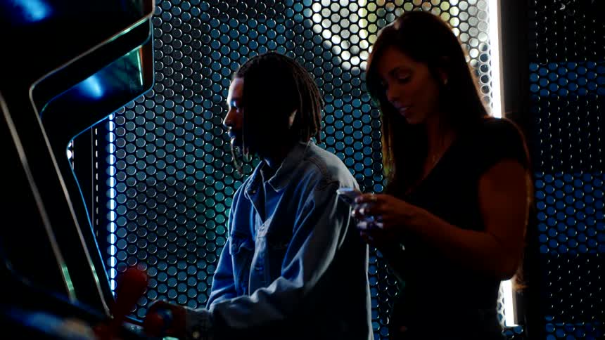 Two people stand at an arcade machine one playing while the other looks at a phone