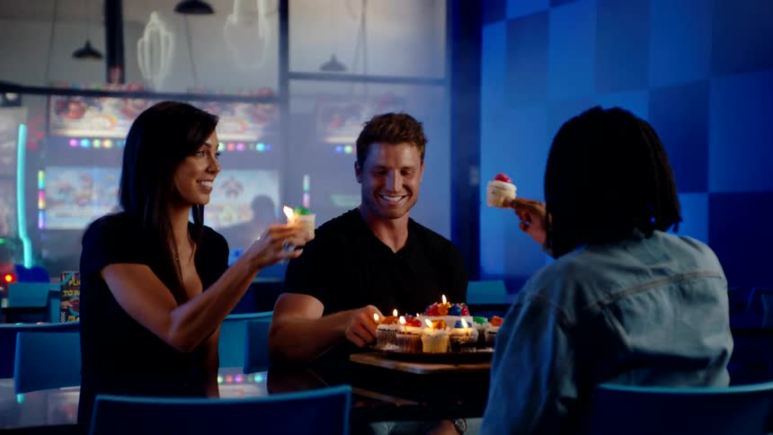 Three friends toast with cupcakes over a lit birthday cake in a vibrant bluelit setting