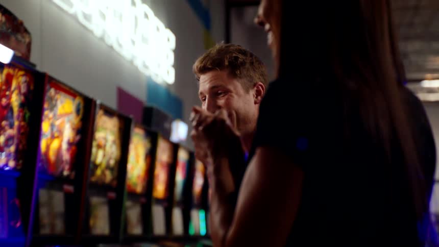 A man and woman enthusiastically play pinball machines in a lively arcade
