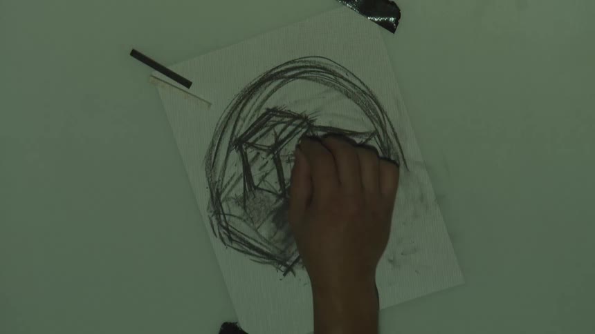 A hand sketches a rough abstract face using dark strokes on paper under lighting