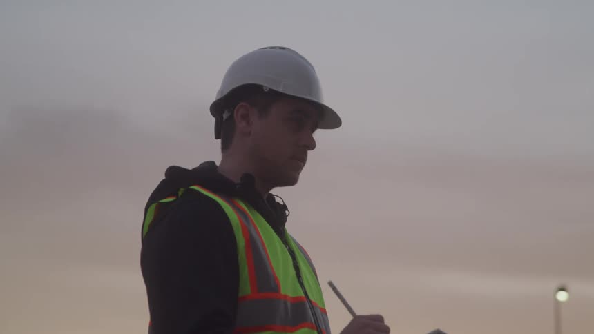 A construction worker in a safety vest and hard hat stands outdoors at dusk taking notes on a clipboard