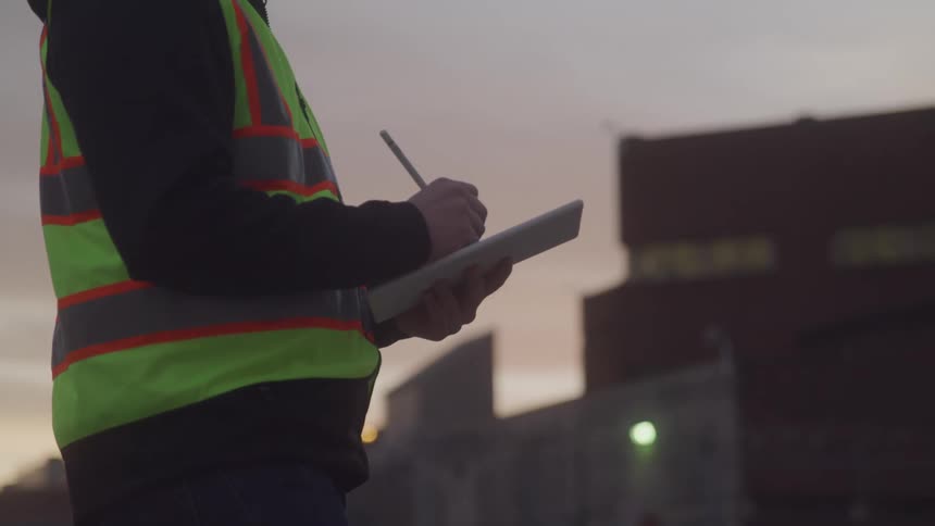 A construction worker writes on a tablet at dusk outside an industrial site