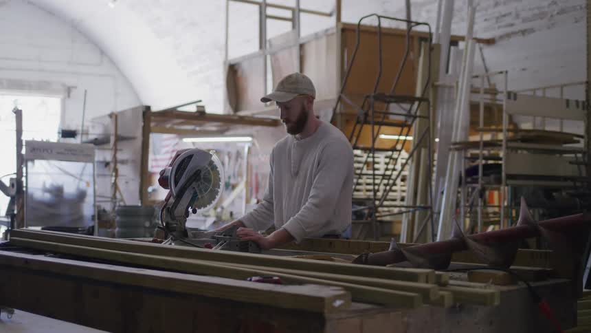 A man operates a miter saw in a cluttered workshop