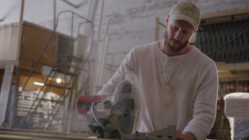 Man uses an electric table saw to cut a piece of wood while standing in a workshop