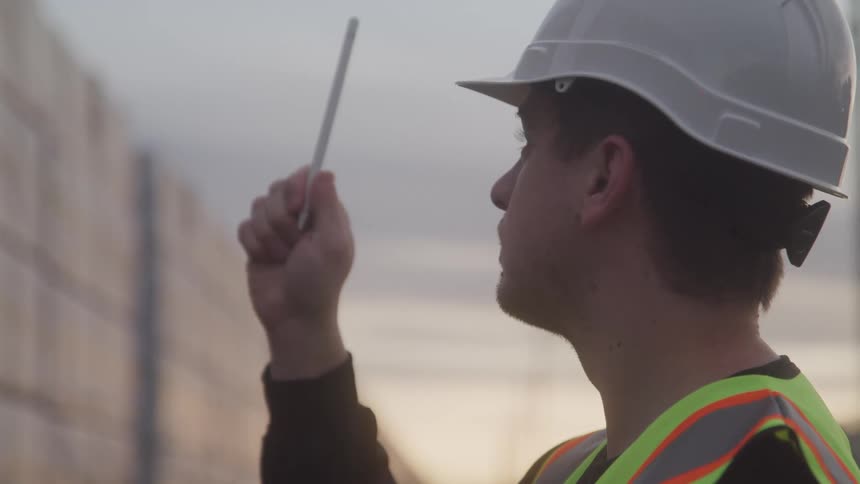 A man wearing a yellow vest and a white safety helmet is looking at a wall and pointing his pen and then writing on a tablet