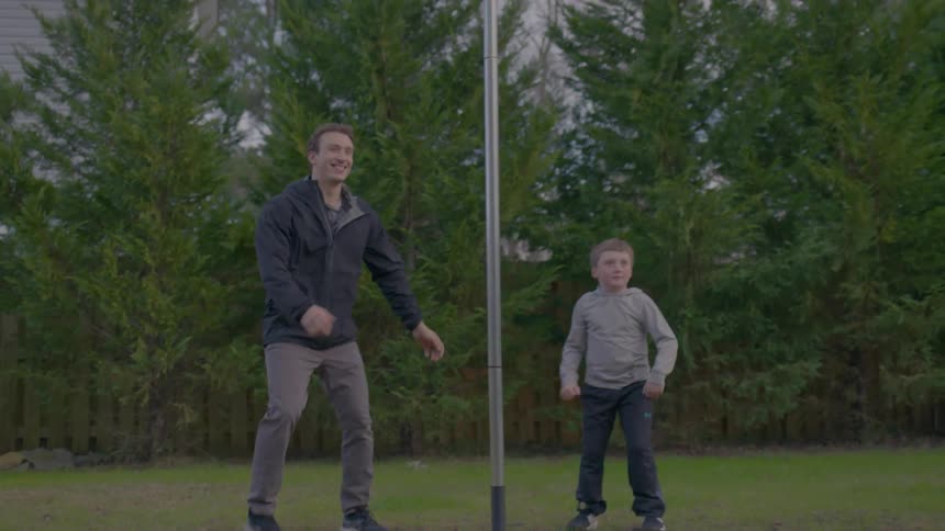 A man hits a tetherball while a young boy watches in a grassy backyard