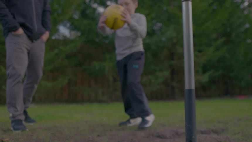A child picks up a tetherball and hits it around the pole as a man stands beside him on a lawn area outdoors