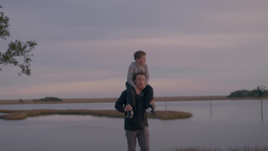 A little boy is sitting on the shoulders of an adult man who are walking and behind them is a large lake