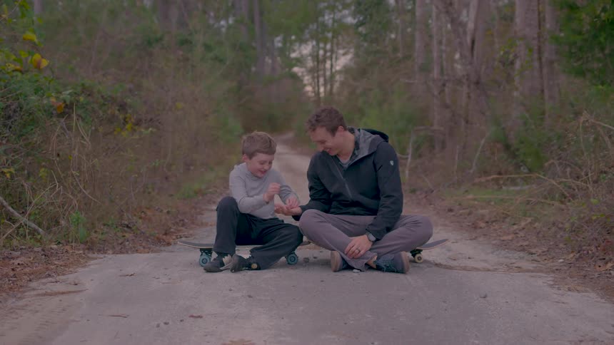 A little boy and an adult man are sitting on a skateboard in an outdoor setting on a road surrounded by trees and vegetation