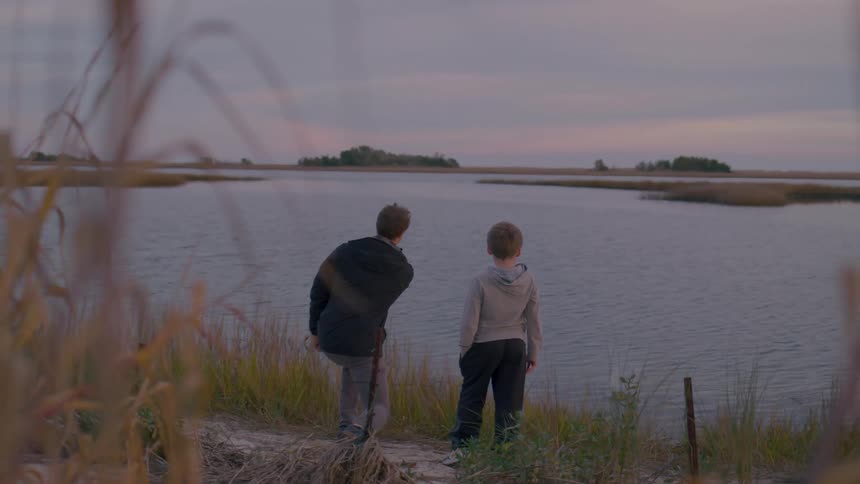 A man and a boy stand by a calm waterfront at dusk gazing at the distant horizon