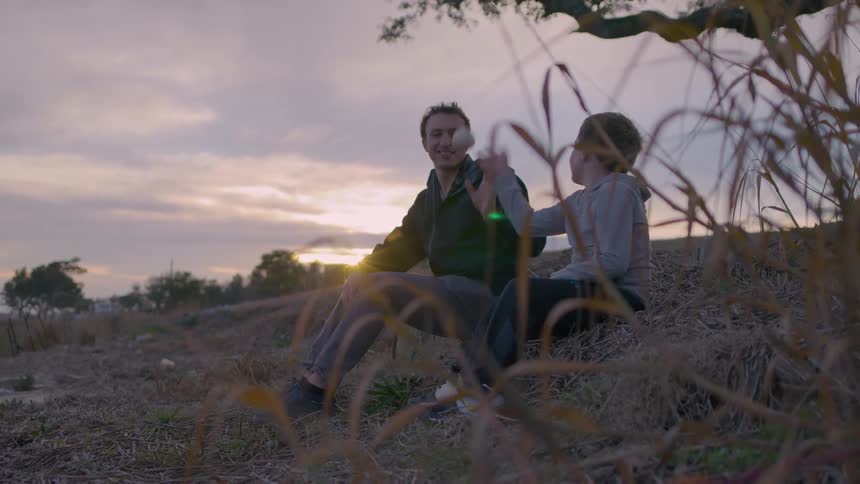 Two people sit outdoors at sunset casually playing catch with a small ball