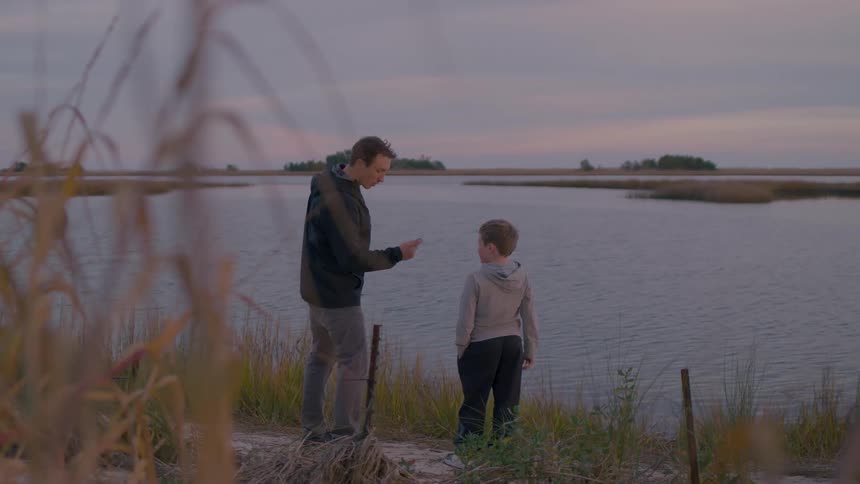 Young man teaches a boy stone skimming as they stand at the river bank at sunset
