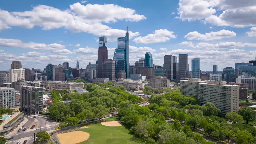 Aerial timelapse view of Philadelphia skyline with green parks in the foreground under a bright sky