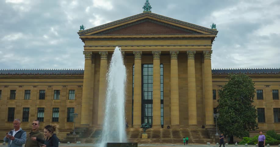 A timelapse captures visitors and tourists moving in front of the grand Philadelphia Museum of Art and its large fountain under an overcast sky