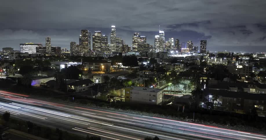 A glowing Los Angeles skyline towers over blurred freeway lights at night
