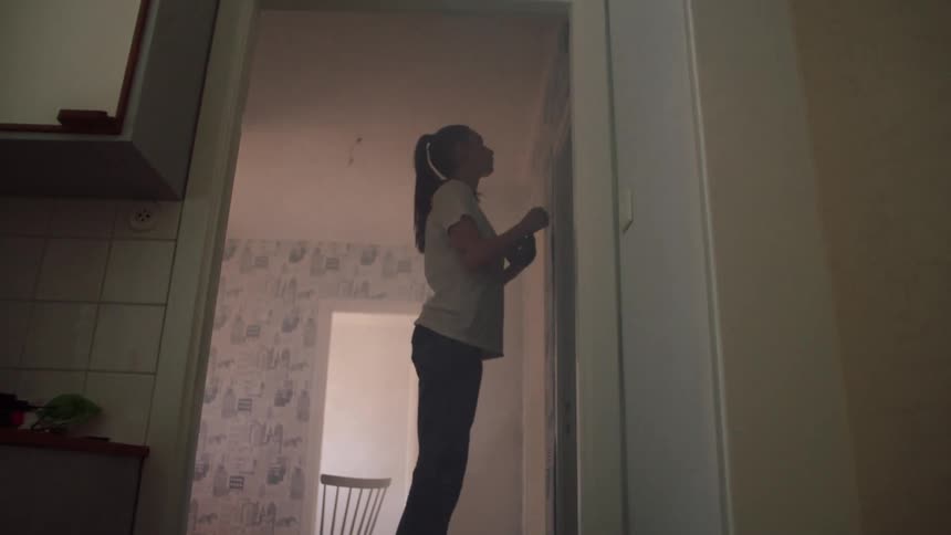 Young woman climbs a metal stool to apply tape at the top area of a room wall