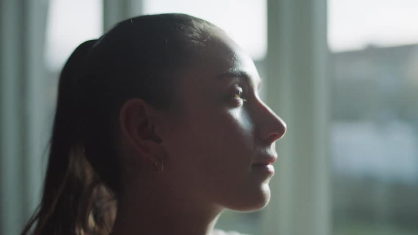 The face of a young woman with ponytail as she is looking forward next to a window