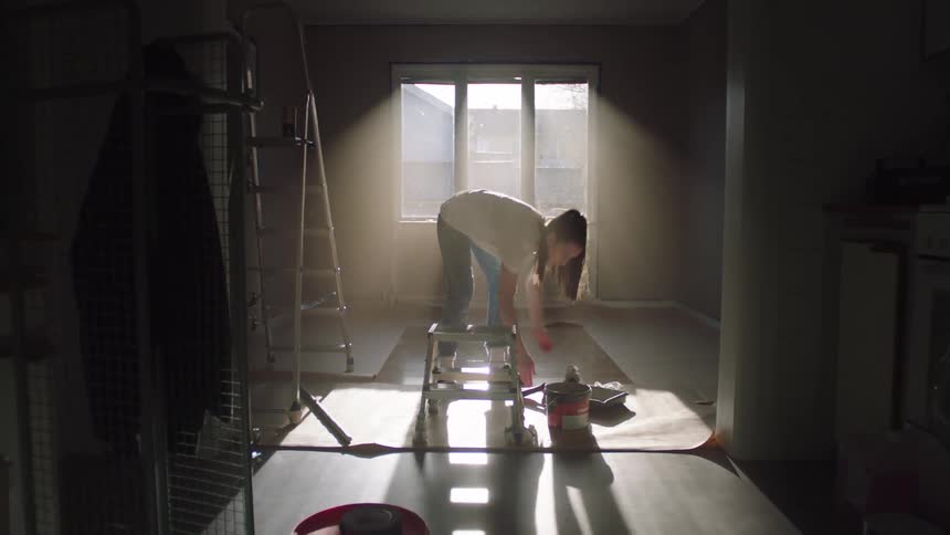 Young woman positions a stool and climbs to paint to top area of a room wall