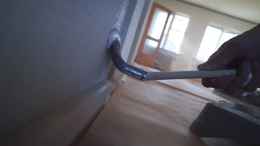 A person spreads white paint with a brush onto a surface in a minimally furnished sunlit room with woodpatterned flooring and scattered construction materials
