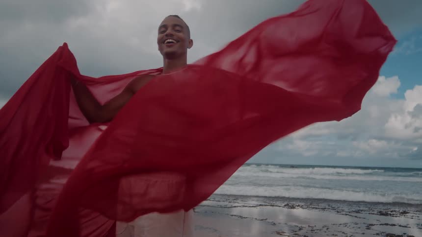A man smiles while holding a red sheet on the beach