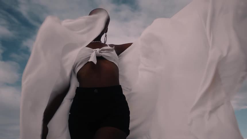 A confident woman poses outdoors with flowing fabric against a dramatic cloudy sky