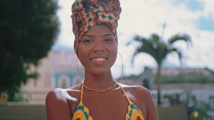 A stylish woman wearing a colorful headwrap and crocheted bikini top smiles confidently in a sun drenched tropical setting