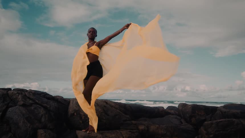 A woman standing on the rocks of a beach and she is holding a yellow piece of clothing that is flying in the wind