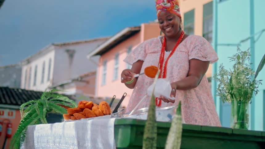 A young black woman serves food onto a piece of paper outdoors around midday
