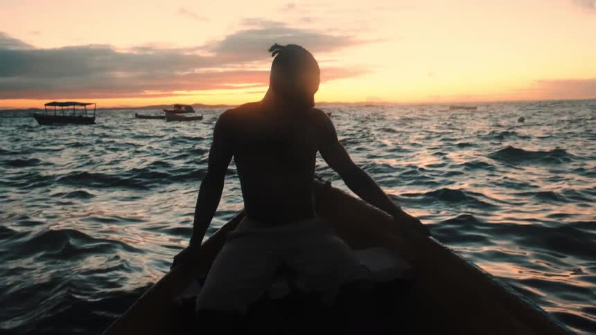 A man sits on the edge of a small boat at sunset silhouetted against the ocean waves