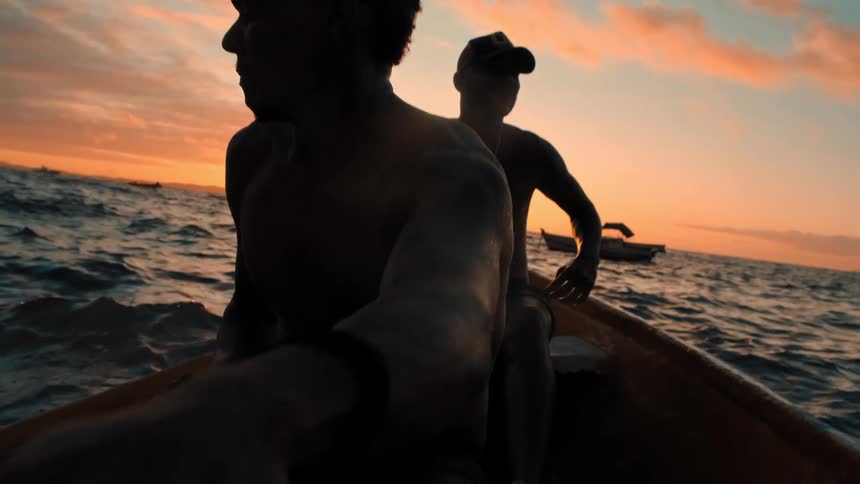 Two people row a boat across calm ocean waters at sunset with another boat visible in the distance