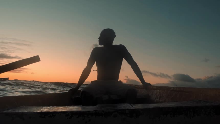 A man is silhouetted as he sits in a boat on the open sea taking a break from rowing against a beautiful sunset