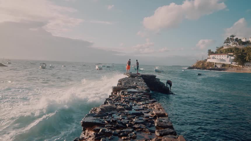 People are standing on a rocky pier extending into the ocean with waves crashing on both sides