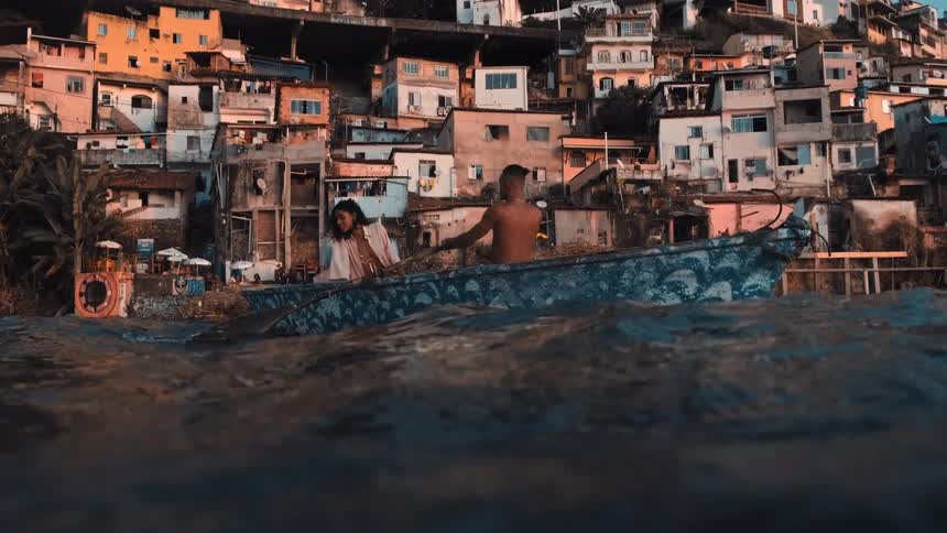 A couple sit on a wooden boat on gentle coastal waters passing a steep seaside village with weathered colorful buildings