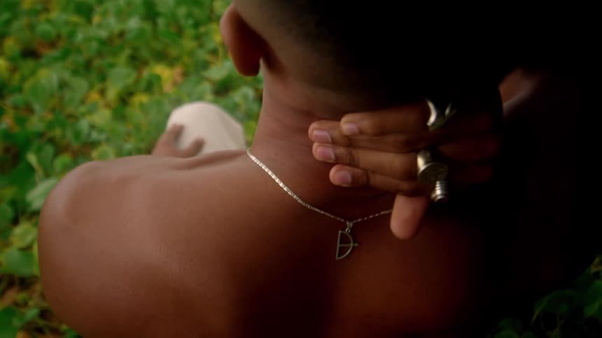 A young man sits on a grassland wearing rings on his fingers and a necklace scratching the back of his head