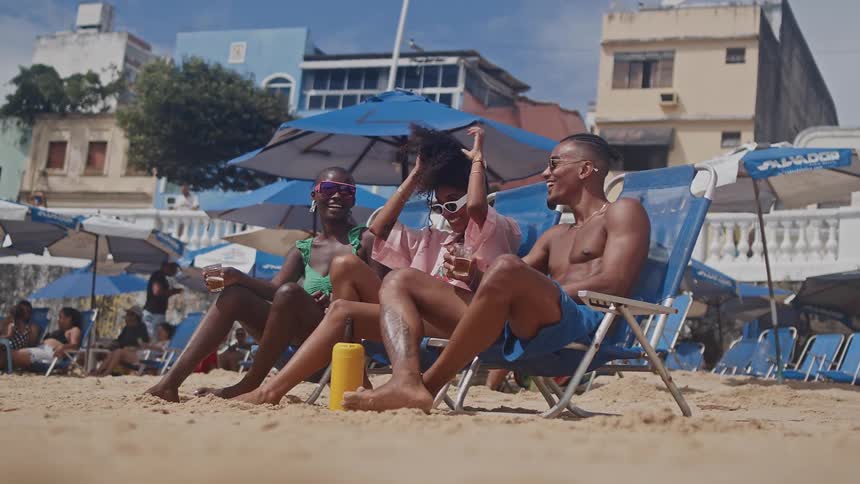 Three friends relax on beach chairs under umbrellas smiling and enjoying drinks on a sunny day