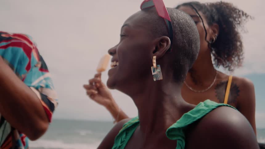 A woman in green ruffled top laughs joyfully surrounded by friends in colorful outfits on a sunny beach