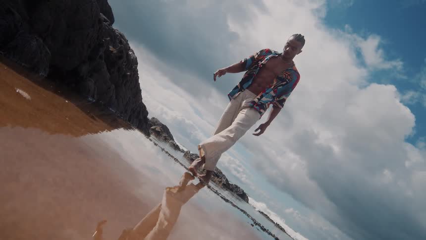 A person walks across a reflective wet beach near rocky outcrops under a cloudy sky