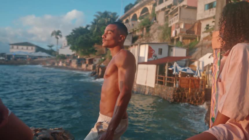A young man stands beside two women near clear turquoise water with a coastal town of palm trees and hillside buildings in the background