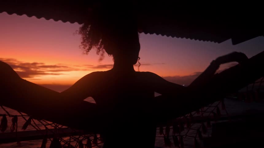 Silhouette of a woman standing leaned back with arms draped over a hammock playing with her spiral curled hair on a porch by the ocean in front of a pink and orange sky background