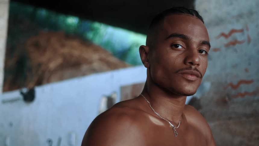A mans face wearing a necklace and he is shirtless and is indoors looking straight ahead