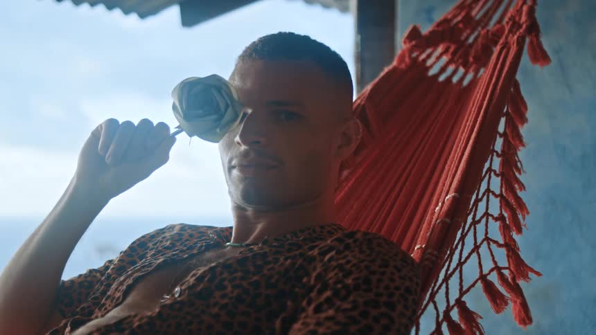 Man on a red hammock in a tropical area holding a white rose over his right eye