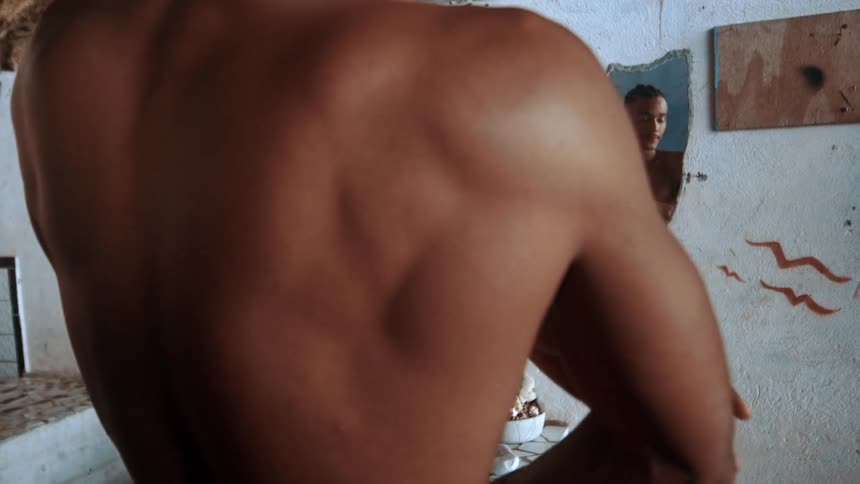 Close up of a black man applying lotion all over his body in front of a wall mirror indoors during daytime