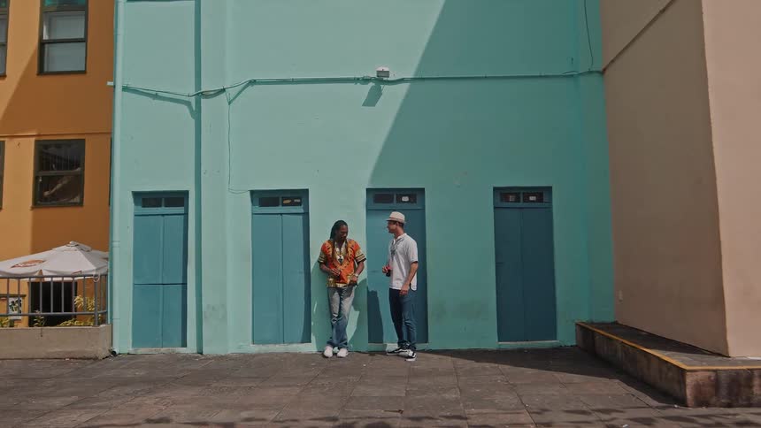 Black man and a caucasian man converse while standing outdoors in front of a blue building during daytime