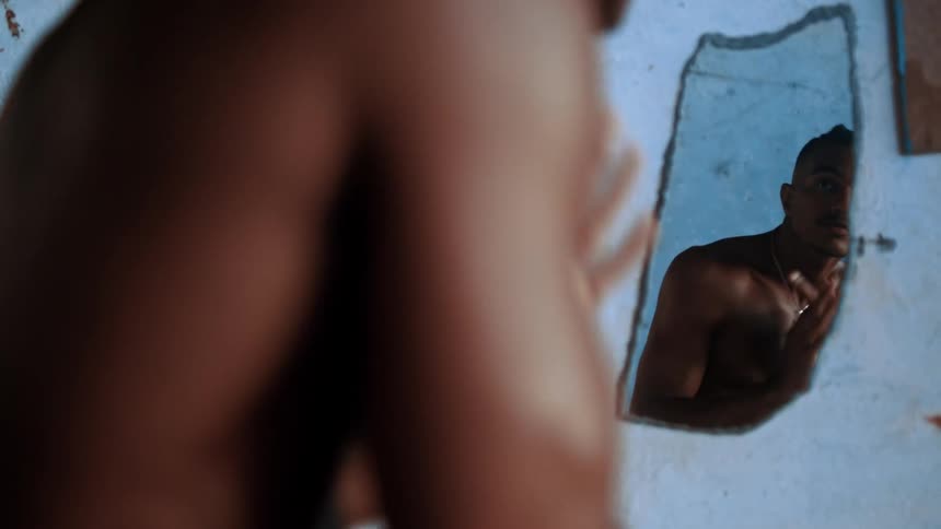 Shirtless man grooming himself in front of a small mirror