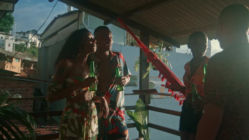 Four peopletwo males and two femalesstand under a shed at a resort midday each holding a bottle of beer laughing and enjoying a lively party atmosphere together