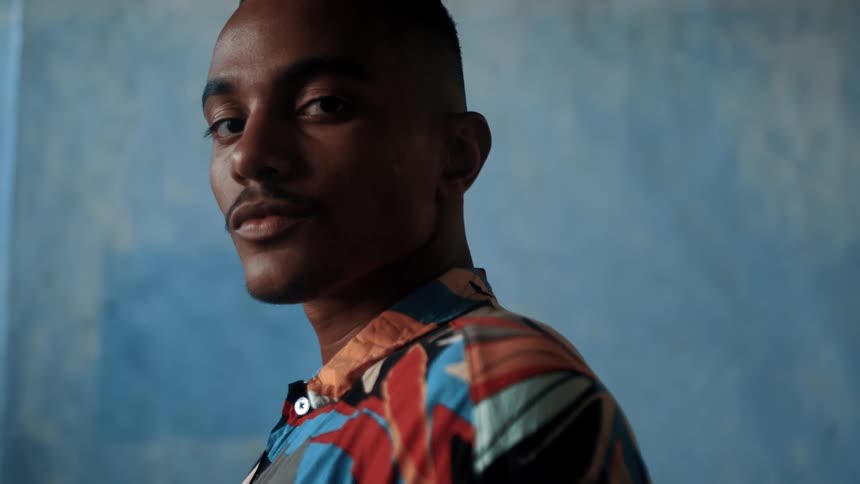 Close up portrait of a young black man standing against a blue coloured wall during daytime
