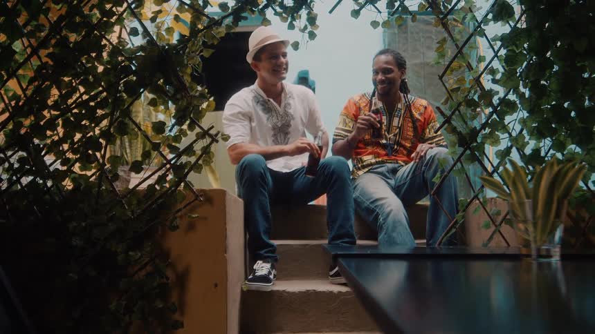 Two men sit on outdoor steps smiling and doing a friendly cheers while enjoying drinks