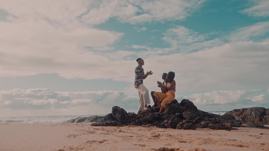 Three friends sit on rocks at the beach and clap their hands