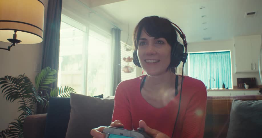 A woman sitting on a couch and wearing headphones is holding a controller while playing a video game and she is happy and makes a celebratory sign with her hand